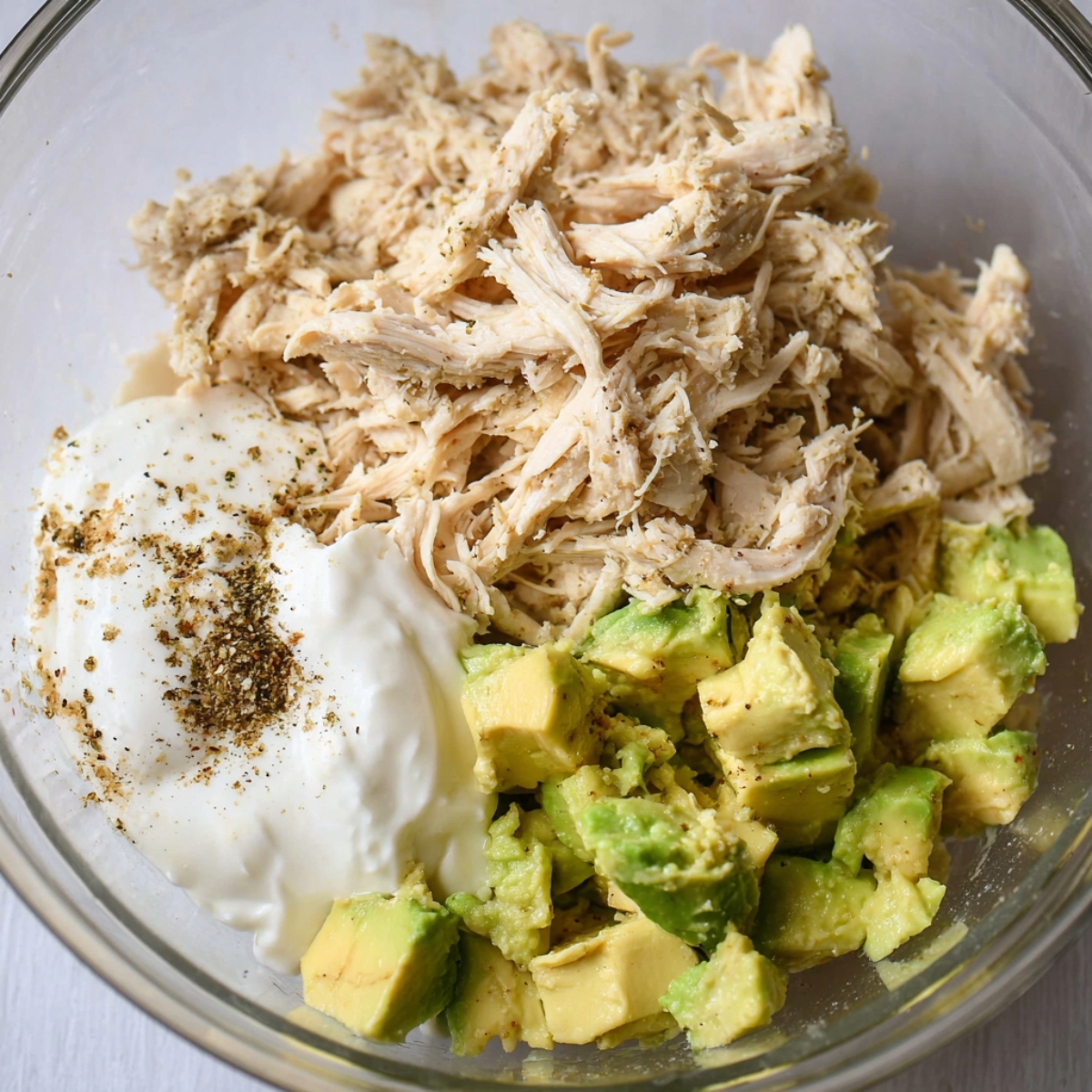 A bowl filled with shredded chicken, diced avocado, yogurt, and spices, being mixed to create a creamy, flavorful filling for wraps.