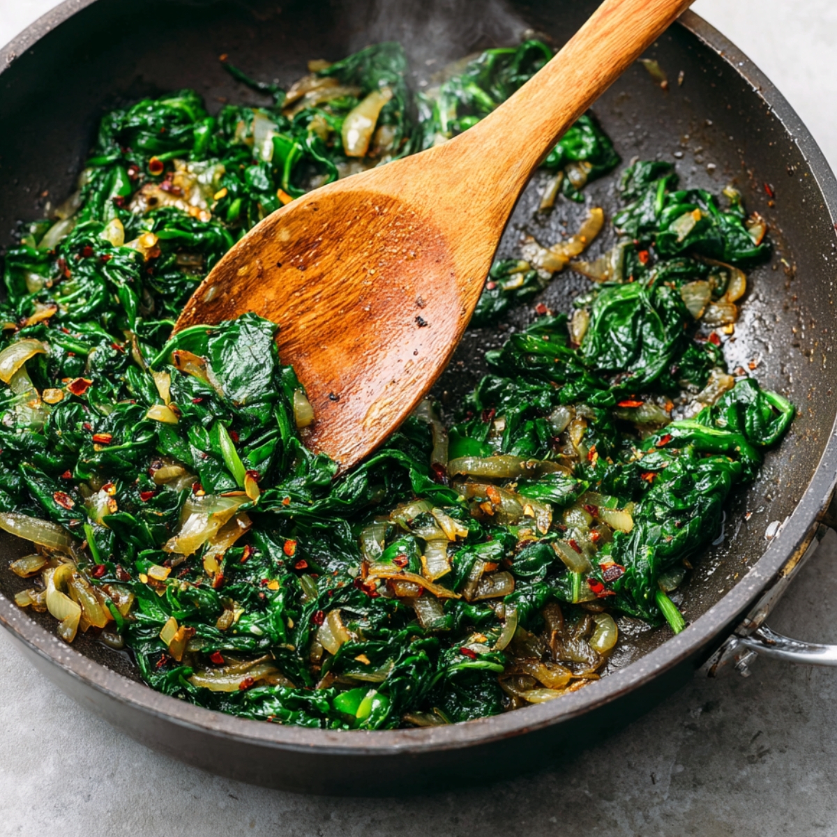Sautéed spinach with onions and red pepper flakes being stirred in a skillet.